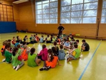 Grundschüler sitzen auf dem Boden der Turnhalle und lauschen dem Handballtrainer